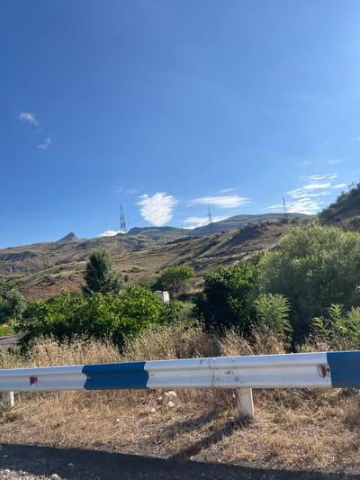 A guardrail with blue and white markings is in the foreground of the image. Behind it, dry, yellow grass covers the ground. Further back, a landscape of rolling hills is visible, dotted with trees and vegetation. Several power line towers are situated on the hillsides, and a distinctive, pointed mountain peak rises in the distance. The sky above is a clear blue with a few wispy white clouds. The scene appears to be a rural or mountainous area in Armenia.