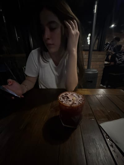 The image depicts a low-light scene inside a restaurant or bar establishment in Santiago, Mexico. A single individual, appearing to be a young adult with long dark hair, is seated at a dark wooden slatted table in the foreground. This person wears a white t-shirt and looks downwards and to their left. Their right hand is positioned near their head, with fingers touching their hair. Their left hand holds a dark-colored smartphone with an illuminated screen; a light-colored bracelet is visible on their left wrist, and their fingernails are painted red.

On the table, slightly to the right of the center, rests a glass containing a dark red beverage with ice and a light-colored garnish, possibly a lemon slice. The rim of the glass is coated with a reddish, granular substance. A dark coaster is beneath the glass. To the far right on the table, a folded white napkin or stack of coasters is present.

In the mid-ground, a tall, dark-colored outdoor patio heater is visible behind the seated individual, adjacent to a brick wall. Further in the blurred background, indistinct figures of other patrons are seated at tables. One background individual on the right wears a dark shirt with a white graphic design on the back. The overall lighting is dim, casting deep shadows.