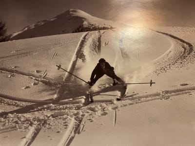 The black and white photograph captures a skier in action on a snowy mountain slope. The skier, a person wearing dark clothing, is in a dynamic pose, leaning into a turn and kicking up a cloud of snow. Their skis are visible, and they are holding ski poles, one of which is angled across their body.
The setting is a vast, snow-covered mountain landscape. In the background, a snow-capped mountain rises under a bright sky, suggesting a clear day, likely in winter. The slope is marked with ski tracks, indicating it's a prepared piste or a popular area for skiing. The lighting suggests it could be daytime, with the sun possibly casting long shadows, although the strong highlight in the upper right corner suggests it might be late afternoon or a specific lighting condition.
The activity is clearly skiing, with the skier exhibiting a skillful maneuver, possibly carving a turn. The spray of snow emphasizes the speed and force of the action. The overall mood conveyed is one of athleticism and exhilaration, capturing a moment of intense physical activity in a natural, winter environment. There is no visible text in the image. The location context "Ленинское, Russia" is not directly discernible from the visual elements of the photograph itself.