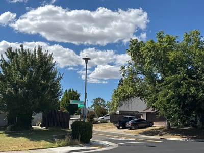This is a daytime, eye-level, exterior shot of a residential intersection in Citrus Heights, California, USA. The scene is dominated by a bright blue sky with scattered white cumulus clouds. A street light stands near the center of the frame, topped with a simple, modern lamp. Two green street signs are attached to the light pole; one reads "Sandridge Street" and the other appears to be "Sunrise Vista".

In the foreground, a paved road curves to the right, with yellow and white lane markings visible. A sidewalk and curb line the edge of the road. To the left, a well-maintained lawn slopes gently upwards towards a dark wooden fence. A large, leafy green tree stands prominently on the left side of the frame, casting shadows on the lawn.

Further back, a residential house with a grey roof and siding is visible. A dark brown garage door is situated on the right side of the house. Several vehicles are parked in the driveway, including a dark grey sedan and a teal pickup truck. Other trees and shrubs are scattered throughout the landscape, adding to the suburban feel of the environment. The overall impression is one of a quiet, sunny afternoon in a suburban neighborhood.