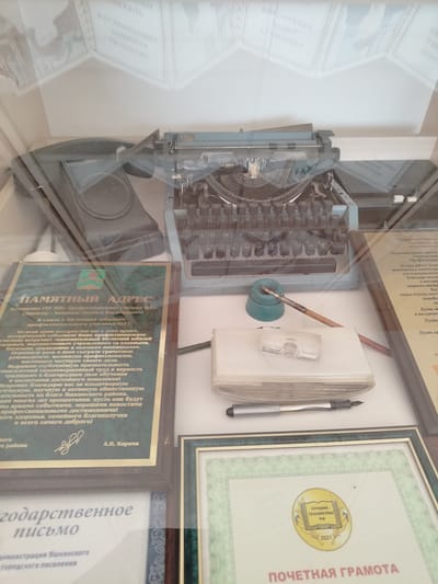 The image shows a glass display case containing several items that appear to be historical artifacts or memorabilia. In the upper half of the display, a vintage blue typewriter is prominently featured, with a sheet of paper partially inserted into it. To the left of the typewriter, a black object, possibly a phone or some other electronic device, is visible. Below the typewriter, on a white surface, a pen and an inkwell are arranged, suggesting a writing or historical documentation theme.
The bottom half of the display contains framed documents. The leftmost framed document is a certificate or award with Russian text and a green emblem. The text includes "Памятный адрес" (Memorial Address) and mentions "Коллективу ГОУ НПО «Профессиональное училище №51» (Директор – Акашкина Людмила Владимировна)" (To the Collective of the State Educational Institution of Vocational Education "Vocational School No. 51" (Director - Lyudmila Vladimirovna Akshina)). The address is dated and likely related to an anniversary or achievement of the vocational school. Below this, another framed document, a "государственное письмо" (state letter), is partially visible, with text about the administration of Yashkinsky district.
To the right, another framed document, a "Почетная грамота" (Honorary Certificate), is visible. It features a circular emblem with the text "ЛУЧШИЕ ТЕХНИКУМЫ РФ" (Best Technical Schools of the Russian Federation) and the year "2021".
The display case is likely located in Yashkino, Russia, given the mention of "Яшкинского района" (Yashkinsky district) on one of the documents. The environment appears to be an indoor setting, possibly a museum, educational institution, or public building showcasing local history and achievements. There are no people visible in the image. The items are arranged neatly within the display case, and the lighting suggests an artificial light source illuminating the exhibits.