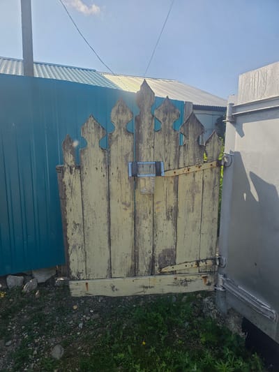 In Georgievka, Russia, an old, weathered yellow wooden gate stands ajar. The gate is adorned with decorative pointed finials along its top edge. Attached to the front of the gate is a clear plastic bottle, secured with tape, suggesting it might be used for collecting something or as a makeshift container. A rusted metal bar crosses the gate horizontally, serving as a decorative element or perhaps reinforcing the structure. The gate is hinged to a grey, industrial-looking structure on the right side of the frame. To the left of the gate, a tall, teal-colored corrugated metal fence stretches into the background. Behind the fence, a house with a silver, corrugated metal roof is partially visible, and a utility pole stands to the left. The ground in front of the gate is covered with a mixture of gravel, rocks, and green grass. The sky above is clear and blue, with a few wispy white clouds. The lighting suggests it is a sunny day.