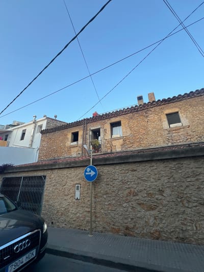The image shows a street scene in L'escala, Spain, under a clear blue sky. The focal point is an old stone building with a tiled roof, featuring several windows. In front of the building, a blue circular sign with a white arrow pointing upwards is visible, indicating a one-way street. Next to the sign, a white rectangular device is mounted on the stone wall.
Below the stone building, a white building with modern features, including stainless steel pipes and windows, is partially visible. A dark-colored Audi car with a license plate reading "7904 HDK" is parked in the foreground on the right. A metal security gate is also present. The scene suggests a sunny day, likely during daylight hours, given the bright sky and shadows. The architecture of the stone building hints at a historical setting, while the modern car and signage indicate a contemporary street. There are no people visible in the image. The overall impression is a quiet street in a Spanish town.