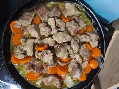 A top-down, close-up shot shows chunks of cooked meat and sliced carrots in a pan. The pan, which appears to be made of dark metal, is filled with a yellowish broth. Some onions are also visible among the meat and carrots. The scene is likely indoors, given the lighting and the presence of a countertop on the right side of the image. The background is blurred but suggests a domestic setting. There is no visible indication of the city of Dolzhikovo, Russia, or any specific geographical markers. The image focuses solely on the food preparation.