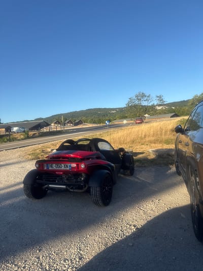 In Charchilla, France, a red three-wheeled vehicle is parked on a gravel surface. The vehicle's license plate reads "HE-050-XN". A brown SUV is parked to the right of the three-wheeled vehicle. In the background, a paved road curves through a dry, grassy field, with several wooden structures and trees lining the landscape. A clear blue sky is visible above.