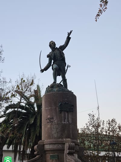 A bronze statue of a man in military attire, with a sword in his left hand and his right arm raised towards the sky, stands atop a stone pedestal in San Fernando, Chile. The statue depicts a historical figure, likely a military leader, in a dynamic pose. The pedestal itself is adorned with inscribed plaques, though their text is not clearly legible in the image. Around the base of the monument, palm trees and deciduous trees with sparse foliage are visible, suggesting a temperate climate. The sky is overcast, casting a soft light on the scene. In the background, a building with a green roof and an antenna can be seen. A street lamp and a sign indicating "Carabineros de Chile" (Chilean Police) are also present in the lower left corner of the image. The overall impression is one of a public monument in a city square.