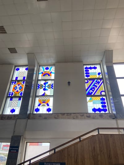 The image shows an interior view of what appears to be a public building, possibly a train station or community center, in Srednii Ikorets, Russia. Three tall, stained-glass windows featuring geometric patterns in blue, orange, and yellow are prominently displayed. The windows are framed by columns with marble accents. The ceiling is a standard white drop ceiling with recessed lighting. Below the windows, there is a band of decorative paneling with a nature-themed pattern, depicting what look like birds. To the left, a sign with Cyrillic text and an arrow indicates "Зал ожидания" (Waiting Room). A set of wooden stairs with a railing is visible in the lower right portion of the image, leading upwards. The overall environment appears somewhat dated, with the building's construction and decor suggesting an older era. The lighting is a mix of natural light from the windows and artificial light from the ceiling fixtures. No individuals are clearly visible in the frame.