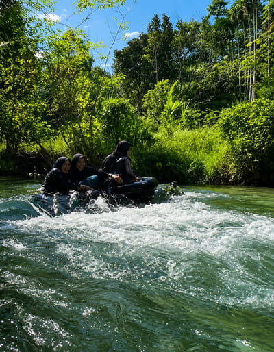 Four individuals, three female and one male, are riding in an inflatable raft on a river. The raft is dark-colored and appears to be moving through choppy water. The three female individuals are wearing dark hijabs and dark clothing. The male individual is to the right of the raft. The raft is positioned in the foreground of the image, with the river flowing rapidly around it. The background consists of lush green vegetation and trees, with a clear blue sky visible above. The scene appears to be set in a natural, tropical environment. This location is identified as Silaiyajae, Indonesia.