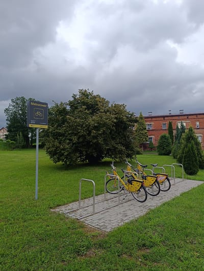 The image captures a scene in Czekanów, Poland, featuring a public bicycle rental station. In the foreground, three yellow rental bicycles are parked in metal racks on a paved area. The bicycles have baskets and branded decals, one of which reads "Sielski Zmieli". To the left of the bicycles, a signpost with the "Metrorower" logo and a bicycle icon is prominently displayed, along with the number "27117" and a phone number. The background includes a lush green lawn, a large deciduous tree, several evergreen trees, and a red brick building. The sky is overcast with dramatic, grey clouds, suggesting inclement weather. The overall environment is an outdoor public space with urban elements.
