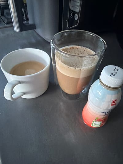 The scene is set indoors on a dark grey countertop in Weert, Netherlands. Three beverage containers are present. To the left, a white ceramic mug with a C-shaped handle contains a light brown liquid, consistent with coffee, exhibiting a thin layer of crema. Centrally, a clear, double-walled glass holds a layered beverage: a darker brown liquid at the bottom, topped by a significant lighter brown, frothy layer. To the right, a translucent, ergonomically shaped plastic bottle with a white screw cap stands upright. The bottle's label features a red-to-light-blue gradient, displaying the "K-Classic" brand logo, the word "PRO," and "Erdbeer" (German for strawberry) alongside an image of strawberries. The bottle cap shows a printed date "10.04" and "42 21128". The background, out of focus, reveals parts of kitchen appliances, including a dark-colored device with visible control buttons, one marked with a power symbol. No individuals are depicted.
