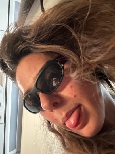 A close-up, angled photograph depicts an individual with light-to-medium skin tone and dark, wavy hair, worn down. The subject is looking towards the viewer, with their head slightly tilted. Dark, wrap-around style sunglasses are worn, obscuring the eyes. A faint, blurred, rectangular reflection, possibly a screen or window, is visible in the right lens (viewer's left). The individual's tongue is partially extended from the mouth. Small dermal imperfections are present on the right cheek (viewer's left). The background is out of focus, showing a vertical white or light-colored appliance or structure on the left side, and a plain, light-colored wall or surface filling the rest of the frame. The scene is indoors. No specific visual cues within the image indicate the location as Sdot Dan Regional Council, Israel.