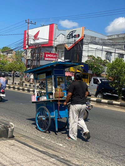 The image depicts a street scene in Sungai Paring, Indonesia, on a sunny day. A man is walking alongside a blue street food cart. The cart is equipped with wheels, a canopy, and appears to be stocked with various items, including bottles, a pink basket, and what looks like a gas cylinder. Behind the cart, a motorcycle is visible. In the background, there are buildings with prominent signage, including a large red and white "HONDA" logo and another sign that reads "MIXUE ICE CREAM & TEA." Power lines crisscross the sky, and several vehicles, including a silver truck and a black car, are parked or driving on the road. Trees and foliage add to the urban environment.