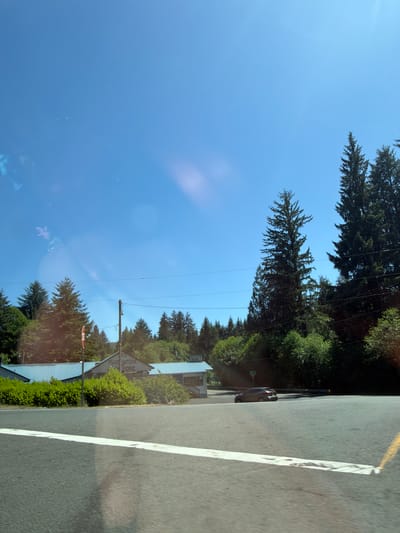 The image, taken from a vehicle's perspective, shows a clear blue sky above a rural or semi-rural setting. In the middle ground, a building with a sign that reads "GAS FOOD STORE" is visible, flanked by green bushes and trees. Another smaller building with a "MARKET" sign is to the right of the food store. A stop sign is positioned to the left of the main building. A dark-colored car is seen driving on the right side of the road, which is in the foreground of the image. Tall pine trees dominate the background, with some smaller trees and vegetation closer to the buildings. The road has a white line marking the edge. The image appears to be taken on a sunny day. While the image depicts a scene consistent with parts of Seaside, Oregon, United States, specific identifying landmarks for Seaside are not definitively present in this frame.