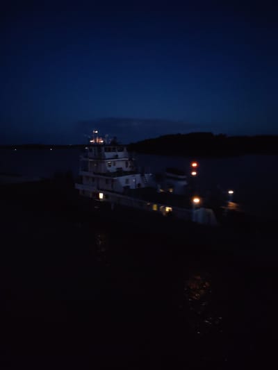 The image shows a large tugboat illuminated by internal and external lights in a dark body of water, presumably at night. The boat is the primary subject, displaying a multi-level superstructure with various light sources visible through windows and on deck. The surrounding environment includes dark land masses along the horizon, suggesting a river or large lake setting. There is no clear evidence of the city of Нова or specific interactions visible within the frame. The overall scene is dark, with the boat's lights providing the main source of illumination.