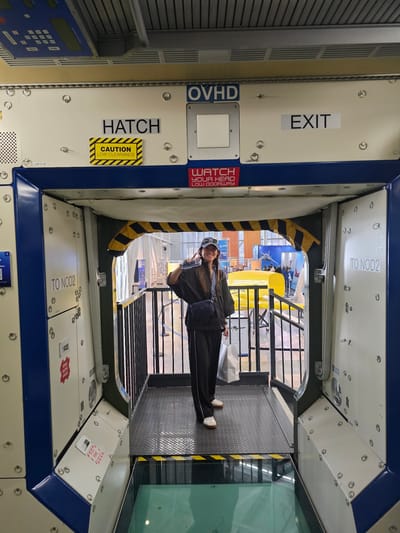 In Mukilteo, Washington, inside what appears to be a museum or exhibition space, a young woman stands within the doorway of a mock spacecraft module. She is wearing a dark tracksuit, a baseball cap, and a crossbody bag, and is holding her phone up, seemingly taking a selfie. The interior of the module is white with blue accents and features text labels such as "OVHD," "HATCH," "EXIT," "TO NOD2," and "CAUTION LOW CLEARANCE." Yellow and black hazard stripes are visible on the ceiling and floor. The woman is positioned on a metal platform, with a checkered floor. Beyond the module, a display area with various exhibits, including yellow structures and what looks like an airplane model, is visible.