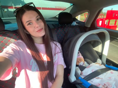 A young woman with long brown hair and sunglasses on her head sits in the front passenger seat of a car, taking a selfie. She is wearing a pink t-shirt with a white graphic design. Next to her, in the back seat, is a baby in a car seat. The baby is wearing a pink hat and has a pacifier in its mouth. The car's interior features black seats with red stitching. Outside the car, through the windows, a red and white sign is visible, as well as two red fire extinguishers and a fence. The scene appears to be at a gas station or service area. The image was taken on a sunny day.