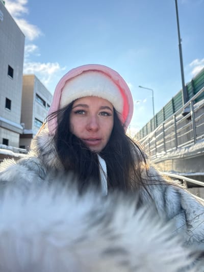 The image presents a close-up, low-angle outdoor portrait of a young adult female in Shul'gino, Russia, taken on a clear, sunny winter day. The subject has long dark hair and is wearing a pink hood with white faux fur lining, consistent with a winter jacket or coat. A light-colored, possibly grey or white, faux fur collar or trim from her outerwear extends into the immediate foreground, creating a soft blur at the bottom of the frame. She gazes directly at the camera with a neutral expression.

In the background, on the left, several multi-story, light-colored urban buildings are visible, showing windows and architectural details. Snow is present on ledges and ground surfaces. On the right, an elevated structure, likely a pedestrian walkway or overpass, features a green sound barrier wall and metal railings, also partially covered in snow. A slender street light pole stands against the bright blue sky, which contains scattered white clouds. The scene is brightly lit by natural sunlight, casting highlights on the snow and structures, and creating a soft glow around the subject.