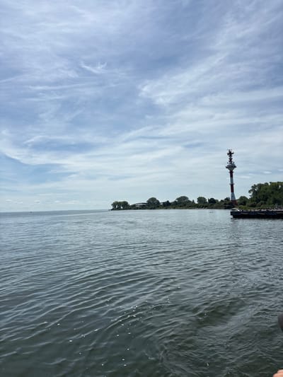 This is an outdoor, eye-level, wide shot taken from the water's surface. In the foreground, the dark, rippling water fills the majority of the frame. In the mid-ground, a calm body of water stretches to the horizon. On the right side of the mid-ground, a long, dark pier juts out into the water. To the left of the pier, a tall, slender lighthouse with red and white stripes stands against a backdrop of green trees and a light blue sky with wispy clouds. In the far distance, a faint bridge structure can be seen. The location is Baltiysk, Russia.