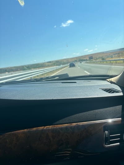 The image is a view from inside a car, looking through the windshield at a highway on a sunny day. The dashboard of the car is visible in the foreground, with a wood-grain trim and an air vent. The highway stretches into the distance, with several lanes and guardrails. Other cars are visible on the highway, and the surrounding landscape appears dry and hilly. There is no specific indication of Vratitsa, Bulgaria, in the image.