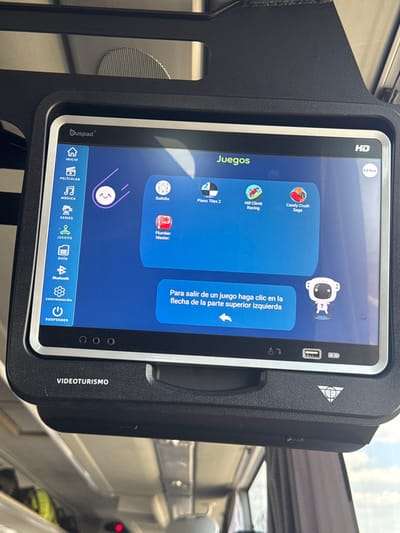 The image shows a tablet-like device with a black frame and silver accents, mounted on a bus or train. The screen displays a menu of games, including Candy Crush Saga, Hill Climb Racing, Piano Tiles 2, Sudoku, and Plumber Master. A cartoon astronaut graphic and a text box with instructions in Spanish are also visible. The bottom of the screen shows navigation icons for Inicio, Películas, Música, Series, Juegos, and Guía, as well as Bluetooth and Susender settings. "VIDEOTURISMO" is printed on the device's side.