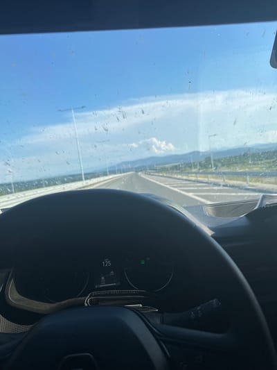 The image is a point-of-view shot from inside a vehicle, looking through the windshield towards a highway. The windshield is visibly dirty with numerous smudges and specks. A steering wheel is in the foreground, obscuring the lower part of the view. The dashboard displays a speedometer showing a speed of 125 km/h.

The highway stretches into the distance, with white lane markings and a barrier on the right. Tall streetlights line the side of the road. The surrounding environment appears to be rural, with green hills and mountains visible in the background under a bright, partly cloudy sky. The time of day appears to be daytime.

The provided context indicates the location is in the city of Dimovo, Bulgaria. While the image itself does not contain specific landmarks or features that definitively place it in Dimovo, it is presented as such.