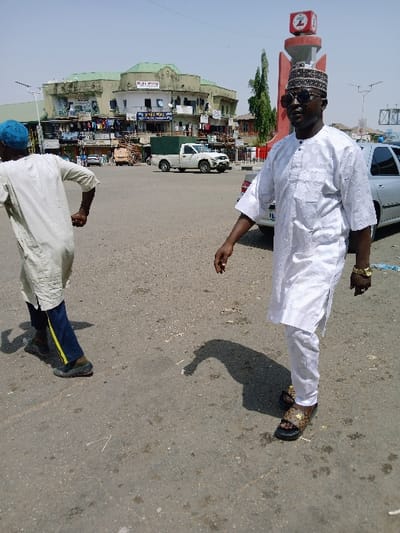 The image depicts an urban street scene in Mumbwa, Zambia, under bright daylight. In the foreground, two adult males are walking on an asphalt surface. On the right, a man is seen walking towards the left of the frame, wearing a white embroidered tunic and trousers, a patterned cap, dark sunglasses, and leopard-print slides. A gold-colored watch is visible on his left wrist. On the left, another man is viewed from behind, walking away towards the left, wearing a light-colored tunic, dark blue trousers with a yellow stripe, and a light blue cap.
In the background, a multi-story commercial building with a light green roof and light-colored walls features multiple storefronts and balconies. Signage on the building includes text such as "DR. M WALA" and "ALMIGHTY." A white pickup truck with a dark green canopy is parked in front of this building. Partially visible on the far right is a silver sedan. A red and white monument-like structure, topped with a symbol resembling a 'Z' or '2', stands behind the man on the right, with a tall green tree adjacent to it. The sky above is clear blue.