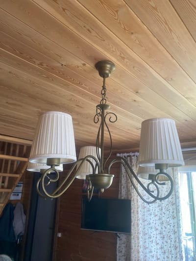 This is a medium-shot, eye-level, indoor, vertical photograph of a rustic-style chandelier with three white pleated fabric lampshades, made of a brass-like material. The chandelier is suspended from a light-colored wooden plank ceiling. To the left of the chandelier, a wooden staircase is visible, leading up to a loft or attic space. Below the chandelier, a dark-colored flat-screen television is mounted on a wooden wall. To the right of the television, sheer white curtains with a floral pattern hang near a window, allowing sunlight to enter the room. The walls are constructed of dark wood planks. The overall impression is that of a cozy, possibly a country house or a dacha in Baryshevo, Russia, given the rustic interior design elements. No people are visible in the frame.