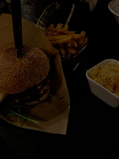 The scene, located in Nowy Targ, Poland, depicts a meal served on a dark, likely black, table within a dimly lit indoor environment. In the foreground, a large burger with a sesame seed bun is positioned on a piece of brown paper, a dark-handled knife inserted vertically through its center. To the immediate right of the burger, a metal wire basket holds a serving of golden-brown french fries. Further to the right, a white rectangular bowl contains a salad composed of shredded cabbage and carrots, consistent with coleslaw or sauerkraut. No individuals are visibly present in the frame.