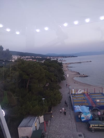 The image captures an aerial view of a coastal scene in Crikvenica, Croatia, likely during the late afternoon or early evening. The foreground is dominated by a paved walkway running alongside a beach. People are seen strolling along this walkway and on the beach, with some gathered on rocky outcrops along the shoreline.
Further down the beach, a children's entertainment area is visible, featuring trampolines and inflatable structures. Adjacent to this is a blue awning with branding that appears to be "Ledo," a Croatian ice cream and frozen food company. A dark-colored car is parked nearby.
The middle ground features a dense line of pine trees, separating the promenade and beach from a town that extends inland. The town consists of white and red-roofed buildings nestled against a backdrop of rolling hills.
In the distance, the Adriatic Sea stretches out to the horizon, with a pier extending into the water. The sky is overcast, creating a soft, diffused light. The top portion of the image shows a reflection, likely from glass, with several circular lights visible.
