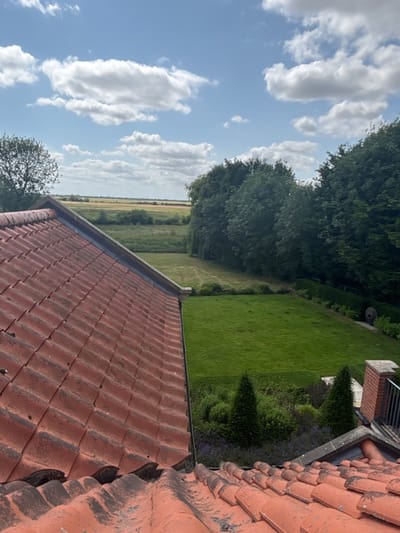 A high-angle shot shows a terra cotta tile roof. Beyond the roof, a view reveals a green lawn, shrubs, and tall trees. Further in the distance, a field stretches towards the horizon beneath a bright, partly cloudy sky. The perspective is slightly tilted, offering a unique view of the landscape.