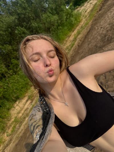 This media file depicts a young woman lying down outdoors, with her eyes closed and her lips puckered in a kiss pose. She is wearing a black tank top and a silver necklace with a small, sparkling pendant. Her blonde hair is spread around her face. The background shows a path or road running through grassy areas, with trees visible in the distance. The scene appears to be in a rural or natural setting. The lighting suggests it is daytime, likely sunny. The woman's pose and facial expression convey a sense of playfulness or affection. There is no visible text in the image. The location context provided is Ryabinino, Russia.
