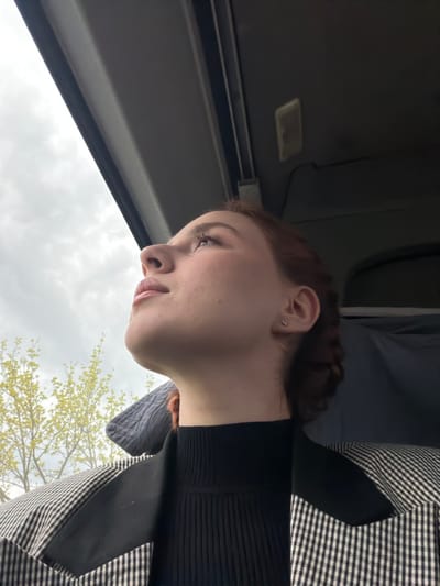 An image depicts a female individual, observed from the mid-chest upwards, looking intently upwards and to the left through a window. Her head is tilted back, with a clear view of her profile. She has fair skin, reddish-brown hair styled in visible braids or twists, and a small silver stud earring in her right ear. Her attire consists of a black ribbed turtleneck top beneath a blazer featuring a black and white checkered pattern and solid black lapels. The individual is situated inside what appears to be a vehicle, indicated by dark grey interior paneling and an overhead light fixture visible in the upper right quadrant of the image. Through the window on the left, an overcast sky is visible, with the upper portion of a tree displaying light green or yellow foliage in the lower left, suggesting an outdoor environment. This scene is located in Eschau, France.