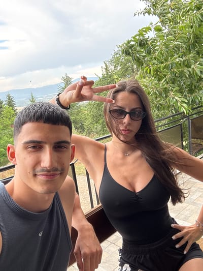 In this selfie, a young couple is posing outdoors. The woman, in the foreground and to the left, is wearing sunglasses, a black bikini top, and black shorts. She is making a peace sign with her left hand. The man, in the foreground and to the right, is wearing a grey tank top. They are both smiling. The background shows lush green trees and a cloudy sky. The setting appears to be a patio or balcony with a tiled floor. The image conveys a relaxed and happy mood, suggesting a casual outing or vacation. The time of day appears to be daytime, with bright, natural light. The location context suggests Vladislavtsi, Bulgaria, which aligns with the natural, green surroundings. There is no visible text in the image.
