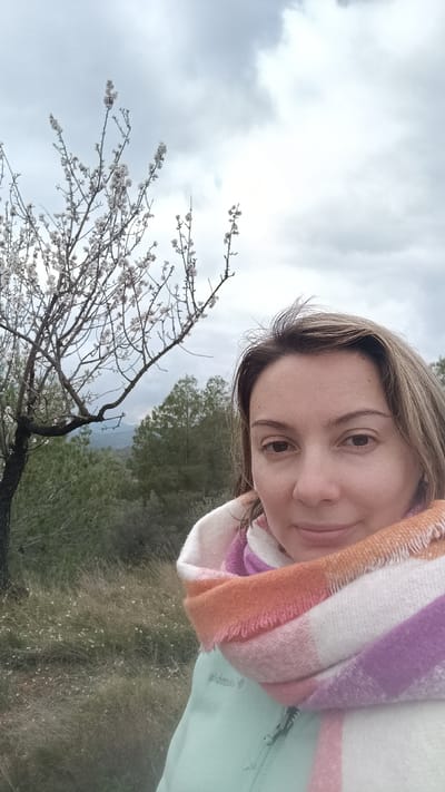 A close-up portrait depicts an adult female positioned outdoors within the city of Jérica, Spain. The subject, a Caucasian woman with light brown hair, occupies the right half of the frame, looking directly into the camera with a subtle smile. She wears a light green zippered jacket and a thick, multi-colored striped scarf featuring shades of white, pink, orange, and purple. To the left, a flowering tree with white blossoms on bare branches is visible, extending into the upper left quadrant. The immediate ground consists of tall, dry grass interspersed with small white flowers. The background features a dense expanse of green trees, likely a forested area, with distant hills or mountains under a heavily overcast, light grey sky.