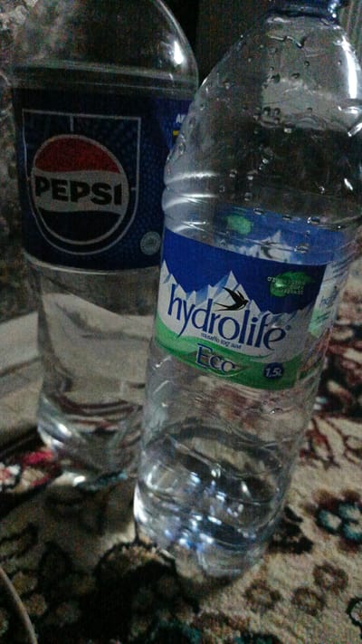 The image shows two plastic bottles of beverages, one labeled "Pepsi" and the other "Hydrolife Eco". Both bottles are clear plastic and appear to be full or partially full of liquid. The Pepsi bottle has a blue label with the iconic red and white Pepsi logo. The Hydrolife bottle has a blue and green label with mountain imagery and the brand name. Both bottles are placed on a patterned surface, possibly a rug or textile. The lighting in the image is dim, creating a slightly grainy effect. The background is out of focus. There is no indication of people or any specific location within the city of Shamsiabad, Uzbekistan.