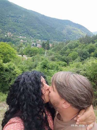 In the foreground, a couple is captured in a close-up shot, embracing and kissing, obscuring their full faces. The woman on the left has long, dark, curly hair and wears a floral-patterned top. The man on the right has medium-length, grayish-brown hair, a beard, and wears a tan t-shirt. The man's left arm is around the woman's neck. In the background, a lush green mountainous landscape is visible, dotted with houses and buildings, indicating a village or town nestled within the hills. The sky above is overcast and white. A date and time stamp, "19.06.2026 15:15", is present in the bottom right corner of the image. The scene appears to be outdoors, in a natural setting, possibly a hillside overlooking a populated area.