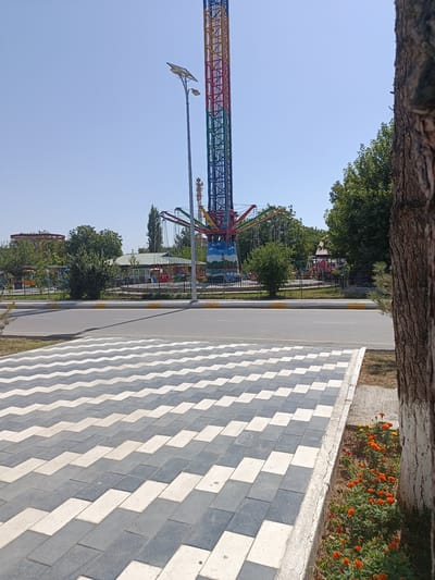 A tall, rainbow-colored amusement park ride, likely a drop tower, dominates the background of this outdoor scene. To its left, a carousel and other carnival-style attractions are visible, some partially obscured by trees. A paved road runs horizontally across the mid-ground, with yellow and white lines marking its edges. In the foreground, a pedestrian walkway with a distinctive zigzag pattern of light gray and white paving stones stretches into the distance. To the far right, the rough bark of a large tree trunk is prominent, partially painted white at its base. Below the tree, a small flowerbed contains vibrant orange marigolds and other red flowers. The sky is clear and blue, suggesting a sunny day. The scene appears to be located in the city of Shahrisabz, Uzbekistan, based on the context provided. There are no people or animals visible in the frame.