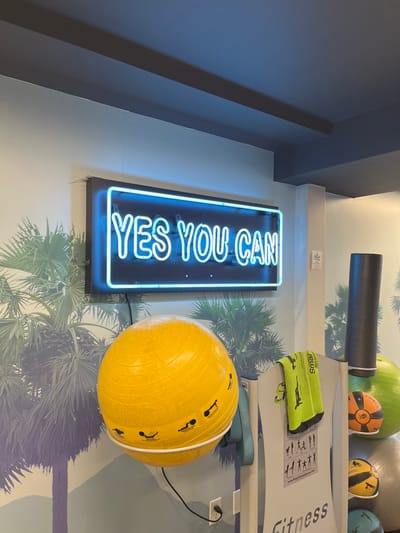 A vertically-oriented neon sign displays the phrase "YES YOU CAN" in bright blue. It is mounted on a wall with a palm tree wallpaper motif. Beside it is a large, yellow exercise ball with stick figure depictions of exercises. Other exercise equipment, including a foam roller and various balls, sit on a shelf in the background.
