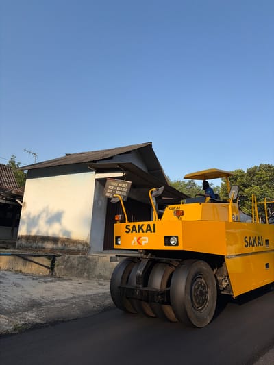 In the city of Kewaluhan, Indonesia, a bright yellow Sakai road roller is actively compacting freshly laid asphalt on a paved road. The roller, bearing the "SAKAI" logo, has a multi-wheeled drum at the front, and a driver is visible in the operator's seat under a yellow canopy. To the left of the roller, a light-colored building with a corrugated metal roof stands under a clear, blue sky. A sign is attached to the building, listing items like "PANDU HELMET," "HELM," "KACA HELM," "MASKER," "SARUNG TANGAN," and "MANTOL." The surrounding environment includes greenery from trees and shadows cast on the building's wall, suggesting it is a sunny day. The scene appears to be in a semi-rural or suburban area, given the building's architecture and the presence of trees.