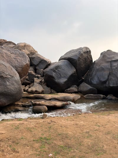 This image depicts a natural rocky landscape in Hale Bandi Harlapura, India, under an overcast sky. The scene is dominated by numerous large, dark-toned boulders, some appearing wet or polished, which are stacked and scattered across the upper and middle ground. A small body of water, possibly a stream or river, flows from the mid-left towards the lower right, creating visible white rapids and foam as it cascades over and around smaller, submerged rocks. The immediate foreground consists of reddish-brown dirt with sparse patches of green grass. A small piece of white and red litter is present on the dirt in the bottom left. No individuals or human-made structures are visible.