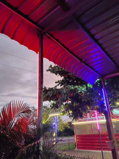 The image depicts an outdoor scene in Dattapur, India, under a covered structure illuminated by red and blue neon lights. In the foreground, large palm leaves with a reddish hue are prominent. Behind them, a tree with green leaves and some red fruits is visible, partially obscuring a red building with a white sign featuring Marathi text. The sign's text translates to "Welcome, we greet you warmly." The building appears to be a roadside establishment. The sky is overcast with soft gray and pink clouds. Electrical wires are strung across the sky. The overall ambiance suggests an evening or twilight setting, likely at a place of business or hospitality.