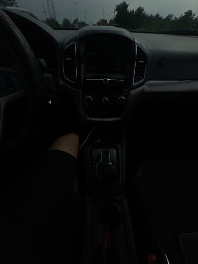 The image shows the interior of a car, likely in the city of Istiqlol, Uzbekistan. The perspective is from the passenger seat, looking towards the dashboard and center console. The dashboard features a prominent infotainment screen and air vents. The gear shifter is visible, with the indicator showing "D" for drive. A person's leg, wearing black attire, is partially visible in the lower left portion of the frame, resting on the passenger side. The environment outside the car, visible through the windshield, appears to be overcast with some trees and what look like power lines or pylons. The lighting is dim, suggesting it could be evening or a very cloudy day. The overall scene is a typical view from inside a vehicle.
