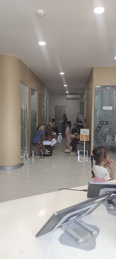 The image depicts a waiting area in what appears to be a clinic or public service building in Keudealue Ie Puteh, Indonesia. The room is well-lit with recessed ceiling lights and a smoke detector is visible on the ceiling. A beige-colored wall runs along the left side of the hallway, with frosted glass partitions. The floor is tiled in a checkerboard pattern of white and grey.
Several people are seated and standing in the hallway, appearing to be waiting. On the left, a woman in a blue hijab and patterned pants is seated, holding papers. Next to her, another woman in a brown hijab is seated. Further down the hall, a group of people are gathered, including a woman in a pink headscarf and a child in a pink outfit. A sign with a wheelchair symbol indicates a disabled-only area. An air conditioning unit is mounted high on the wall. A fire extinguisher is visible next to a green box on the wall.
In the foreground, a white counter is visible with a tablet computer on a stand. A young girl with dark hair in a braid is partially visible in the lower right corner, looking to the right.