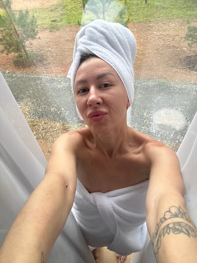 This is a selfie taken by a woman wrapped in a white towel, with a white towel wrapped around her head. She is looking directly at the camera with a slight smile. Her left arm is visible, displaying a tattoo of a floral wreath. She is wearing a white robe that is partially visible. The background shows a blurred outdoor scene with green foliage and gravel. A circular manhole cover with decorative detailing is visible on the ground behind her. White sheer curtains are in the foreground, framing the image. The lighting suggests it is daytime. The overall impression is of a relaxed, possibly post-shower or spa-like moment. There is a tag visible on her robe with the word "HORECA" printed on it.