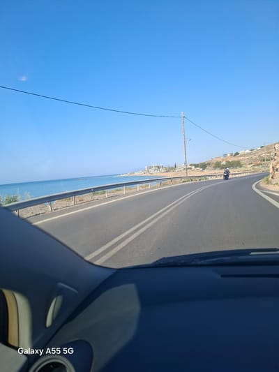 The image, taken from inside a vehicle in the Gouves Municipal Unit, Greece, shows a coastal road running alongside the sea. The road, marked with double white lines, curves slightly. A metal guardrail separates the road from the sea. A motorcycle is visible on the road in the distance. A utility pole stands on the roadside. The sky is clear and blue.