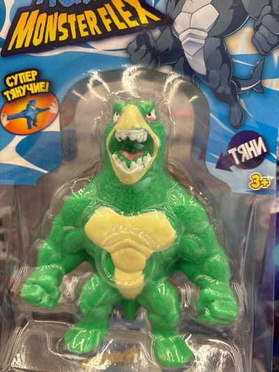 This is a product shot of a "Monster Flex" toy, a green, humanoid-like creature with a yellow chest, in its original packaging. The toy is in the center of the frame, with its mouth wide open, revealing sharp white teeth and a pink tongue. The packaging is clear plastic, allowing the toy to be fully visible. Above the toy, the "Monster Flex" logo is prominently displayed in yellow lettering. To the left of the toy, there is a circular graphic with a blue shark-like creature, and text that appears to be in Russian. To the right of the toy, there is a circular graphic with text and an arrow, also appearing to be in Russian, and a yellow circle with "3+" inside, indicating an age recommendation. The background of the packaging features a blue, wave-like design with an illustration of a large, grey, monster-like creature with fins and a tail. The image does not provide any specific details about the city of Kuzmolovsky, Russia, and therefore cannot be placed in that location.