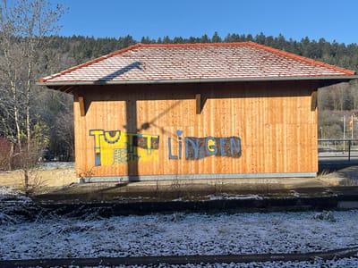 A small, rectangular building with a terracotta-tiled roof dusted with snow. It's constructed of light-colored wooden planks and features graffiti in yellow and blue. The word "TUTTLINGEN" is visible. The structure stands on a concrete base with a foreground of frosty grass and railway tracks. Behind the building, a hillside covered with dense trees rises. The sky is clear and blue.