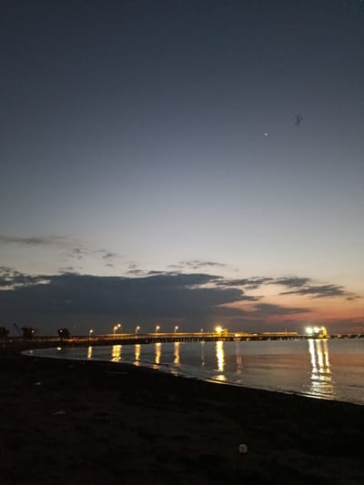 This image depicts a twilight scene at Jangkar, Indonesia, featuring a long pier stretching into the calm sea. The sky transitions from a deep dusk blue at the top to softer hues of grey and hints of orange near the horizon, suggesting either dawn or dusk. Scattered clouds are visible, with darker, more defined clouds present lower in the sky.
The pier is illuminated by a series of evenly spaced streetlights, casting bright yellow glows that reflect vividly on the water's surface. These reflections create elongated, shimmering streaks of light. On the left side of the image, industrial elements, possibly cranes, are faintly visible against the darkening sky.
The foreground consists of a dark, sandy shore, sparsely dotted with white specks that could be debris or small objects. The water appears relatively still, with gentle ripples. The overall mood is peaceful and serene, with the artificial lights of the pier providing a striking contrast to the natural twilight.
There are no visible people or text in the image. The time of day is clearly indicated by the lighting and sky conditions. The location cues (pier, industrial elements, twilight over the sea) are consistent with a coastal area.