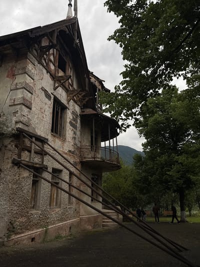 The image depicts a severely dilapidated, multi-story building with crumbling plaster and exposed wooden structural elements. The architecture suggests an older style, possibly an abandoned mansion or historical structure. The facade shows significant decay, with cracked walls, missing plaster, and broken windows. A partial balcony juts out from the upper floor, its railing rusted and in disrepair. A rudimentary wooden support structure, possibly a makeshift staircase or bracing, is attached to the side of the building.

In the background, lush green trees dominate the scene, with a misty, overcast sky above. A mountain range is visible in the distance, suggesting a rural or semi-rural setting. Several individuals are present in the middle ground, appearing to be walking or standing in a park-like area adjacent to the building. Two park benches are visible, one occupied by two individuals, and another empty. The ground in the foreground is dark and appears to be unpaved, possibly a dirt path or graveled area. The overall atmosphere is somber and evokes a sense of abandonment and disuse. The context provided places this scene in Akhtala, Armenia, though specific landmarks within Akhtala are not identifiable from the image alone.