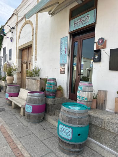 The image shows the exterior of a shop called "KOROS ENOTECA" in Crabonaxa/Villasimius, Italy. The shop has a white facade with a wooden door and a large window. Above the door is a sign that reads "KOROS ENOTECA". In front of the shop, there are several wooden barrels used as decorative elements. Two barrels on the left have a pink band with the shop's logo. The barrel in the center right has "WINE SHOP ENOTECA" painted on it in teal. A third barrel in the foreground also has the shop's logo on a teal band. A stone bench is placed in front of the barrels, and potted plants are visible on the steps leading to the shop. A black mailbox is mounted on the wall to the right of the door. The street in front of the shop is paved with stone tiles.